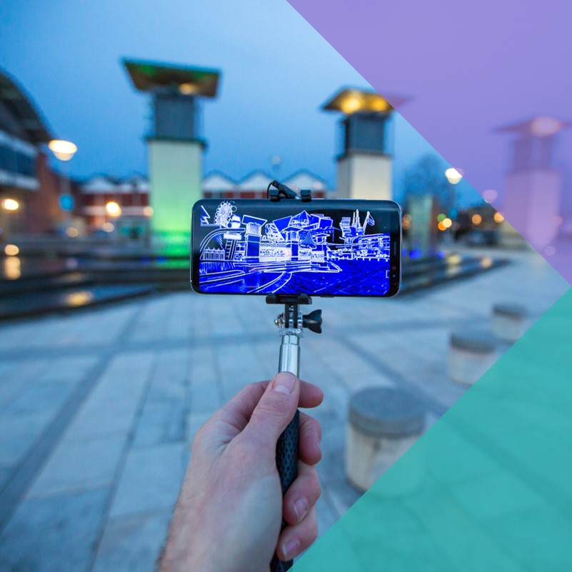 A photograph of a phone held afore the camera on a selfie stick with a AR (augmented-reality) display of the scene in front of them